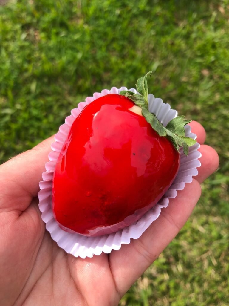 Mão segurando um morango do amor, coberto com calda vermelha brilhante, em uma forminha de papel, com gramado verde ao fundo.