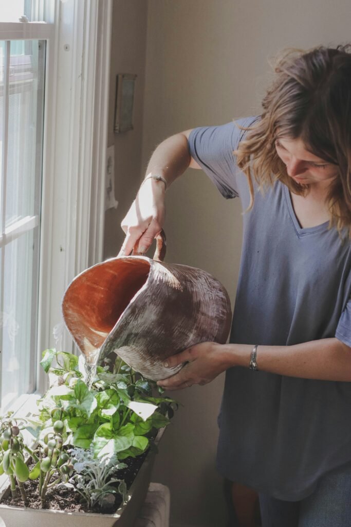 Mulher regando plantas em um vaso retangular próximo a uma janela, usando um regador de cerâmica grande, sob luz natural.