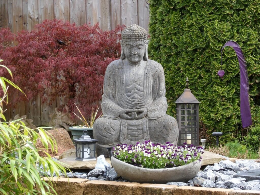 Jardim zen com estátua de Buda ao centro, cercada por flores roxas em vaso de pedra, lanternas decorativas, pedras brancas e plantas ornamentais ao fundo.