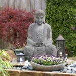 Jardim zen com estátua de Buda ao centro, cercada por flores roxas em vaso de pedra, lanternas decorativas, pedras brancas e plantas ornamentais ao fundo.