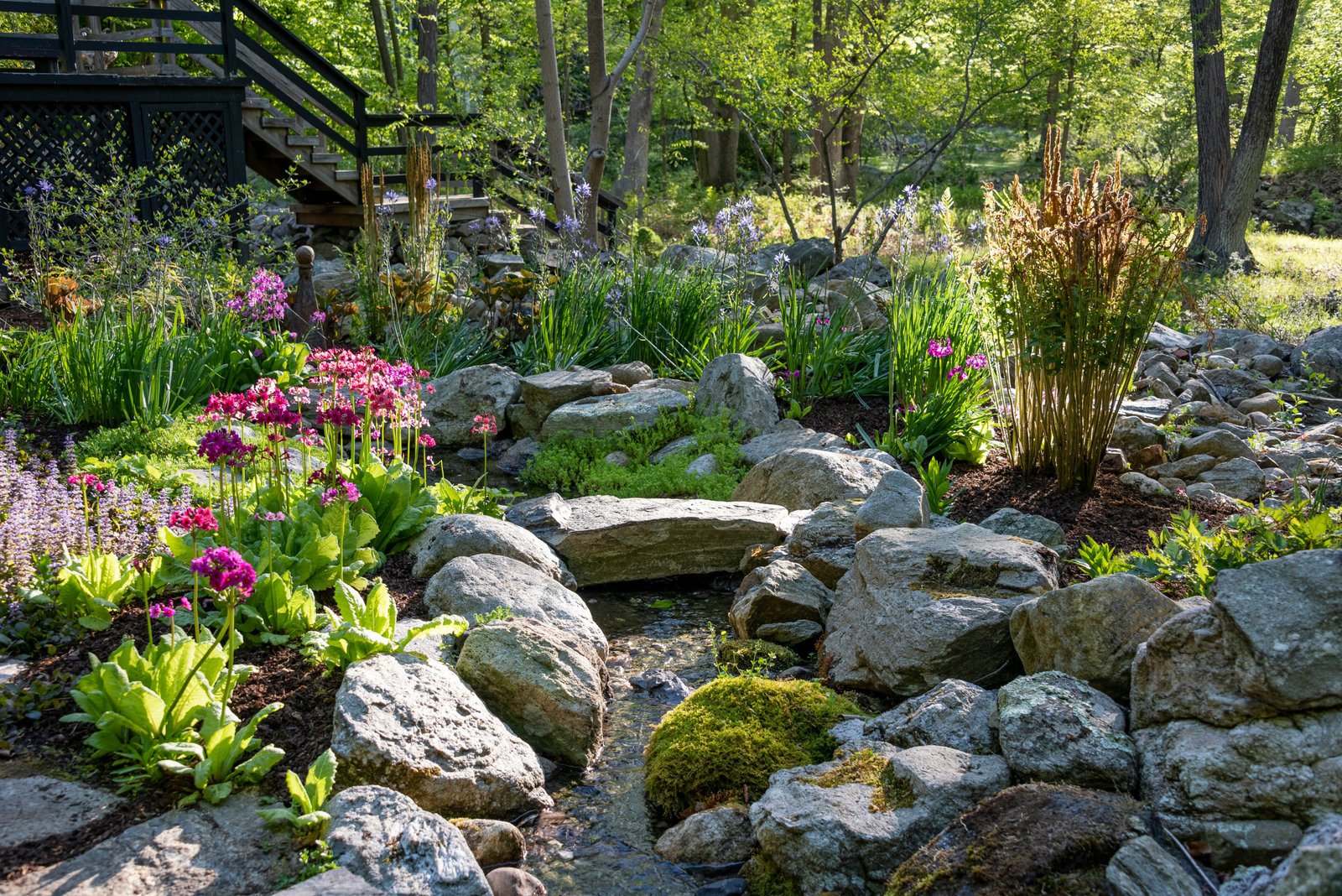 Jardim zen ao ar livre com pedras naturais, pequeno curso de água formando cascata, musgos e flores coloridas, cercado por vegetação e árvores.
