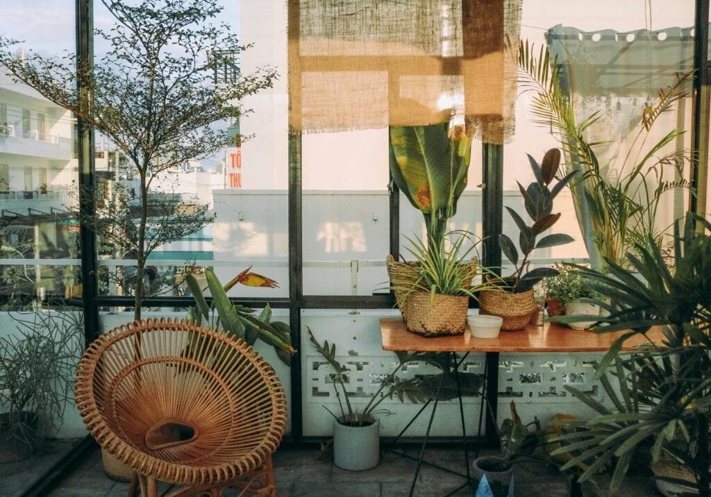 Jardim de inverno para sala em ambiente envidraçado, com várias plantas em vasos e cestos de palha, cadeira de vime e mesa de madeira iluminada pela luz natural.