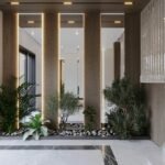Jardim de inverno para sala moderna, com plantas verdes dispostas no chão entre colunas iluminadas, pedras brancas decorativas, piso de mármore e grandes janelas de vidro.