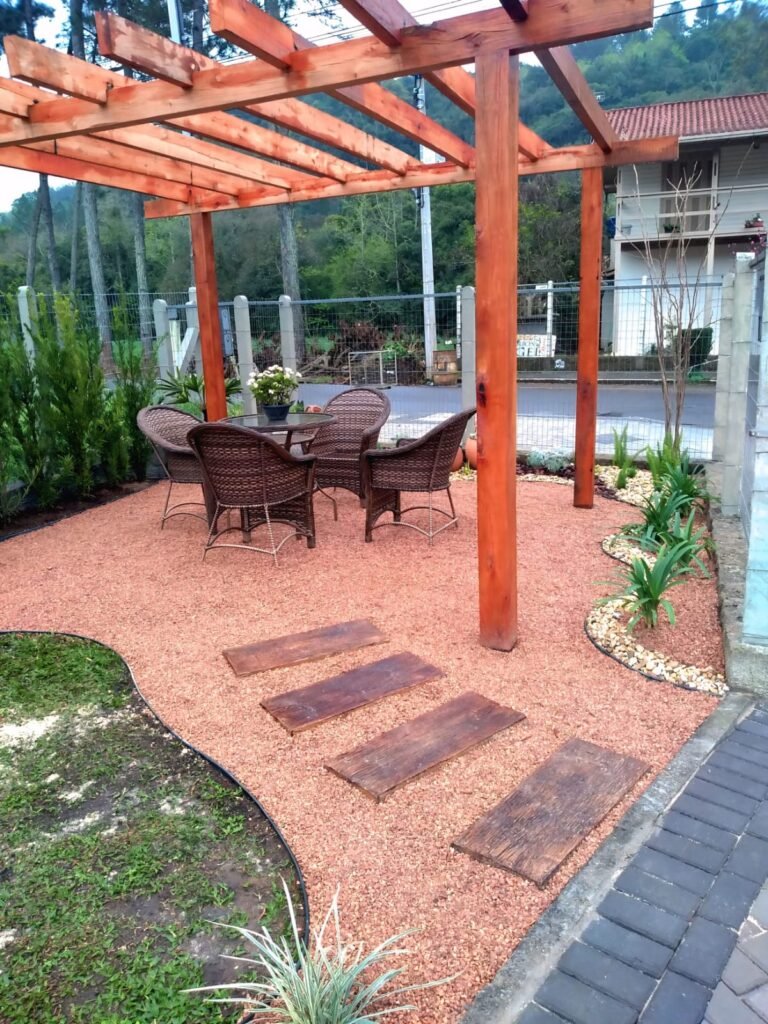 Jardim para quintal pequeno com pergolado de madeira, conjunto de mesa e cadeiras de fibra sintética, caminho de madeira sobre pedrisco e canteiros laterais com plantas e pedras decorativas
