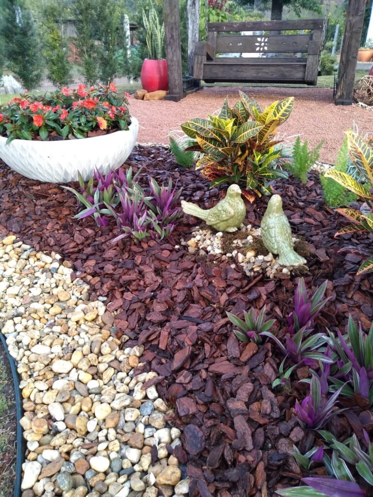 Jardim para quintal pequeno com canteiro de flores coloridas, vasos decorativos, pedras ornamentais, cobertura de casca de pinus, plantas tropicais e esculturas de pássaros de cerâmica