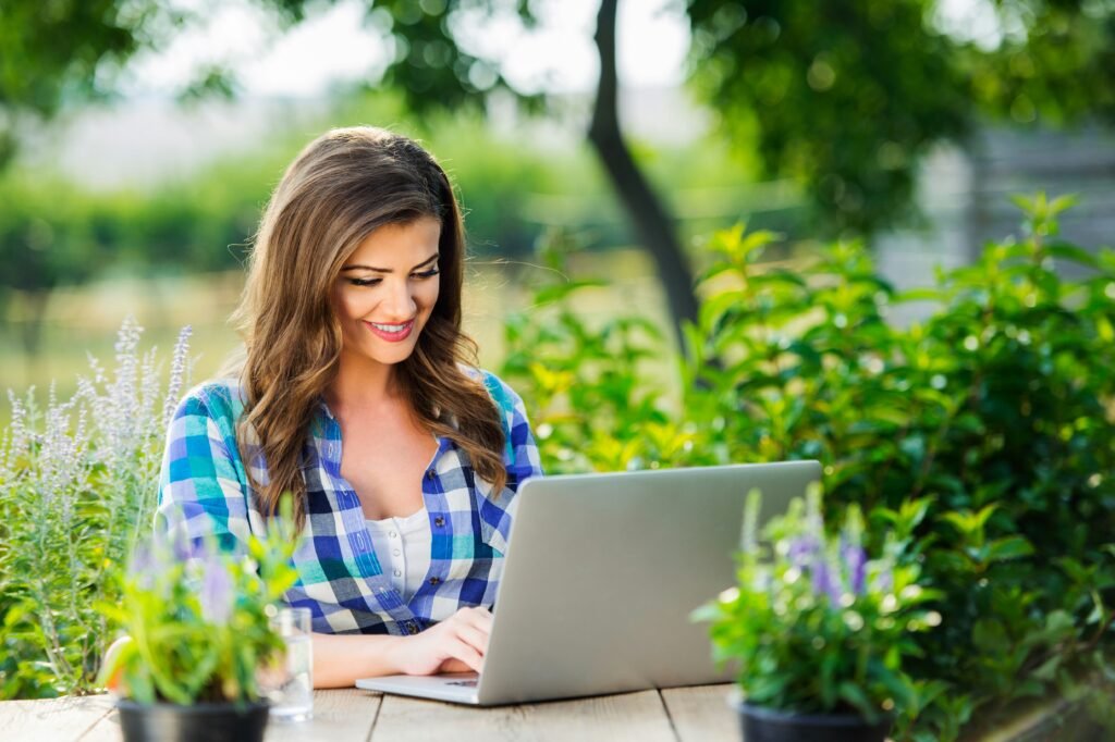 Mulher sorridente usando notebook em meio a plantas verdes sobre a mesa, representando o planejamento e checklist para cuidados de jardinagem.