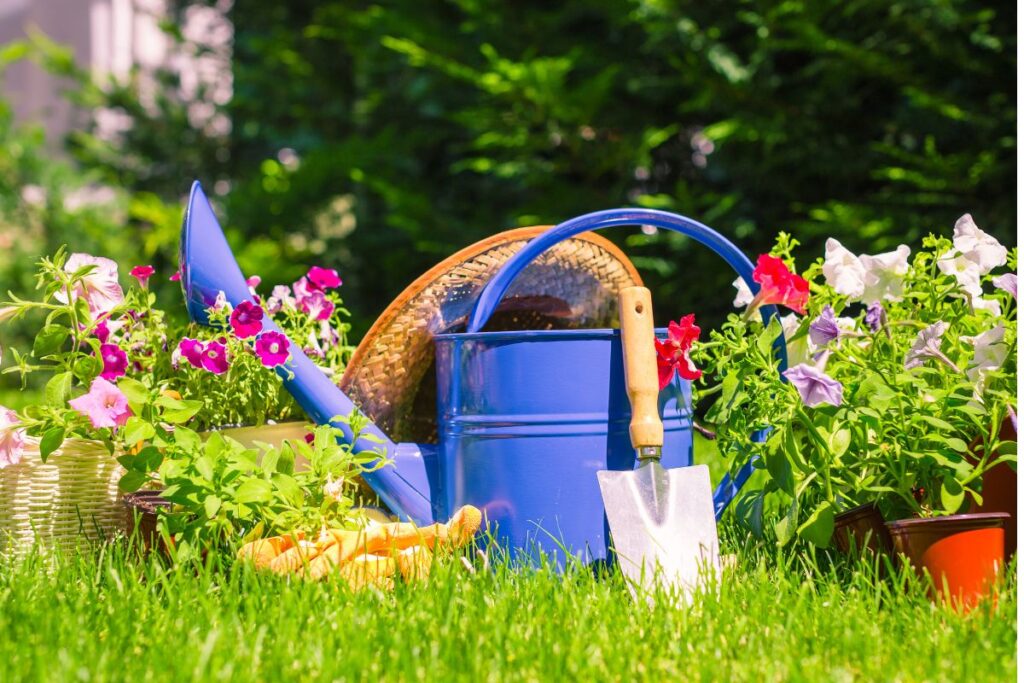 Itens de jardinagem com regador azul, pá e flores coloridas em vasos sobre grama verde, prontos para criar espaços verdes organizados e funcionais.