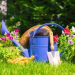 Itens de jardinagem com regador azul, pá e flores coloridas em vasos sobre grama verde, prontos para criar espaços verdes organizados e funcionais.