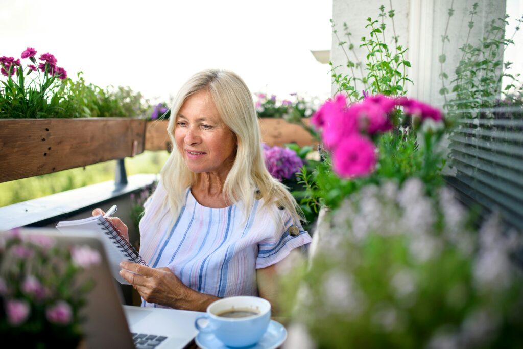 mulher planejando um espaço verde em varanda com flores e plantas ao redor, usando caderno e notebook