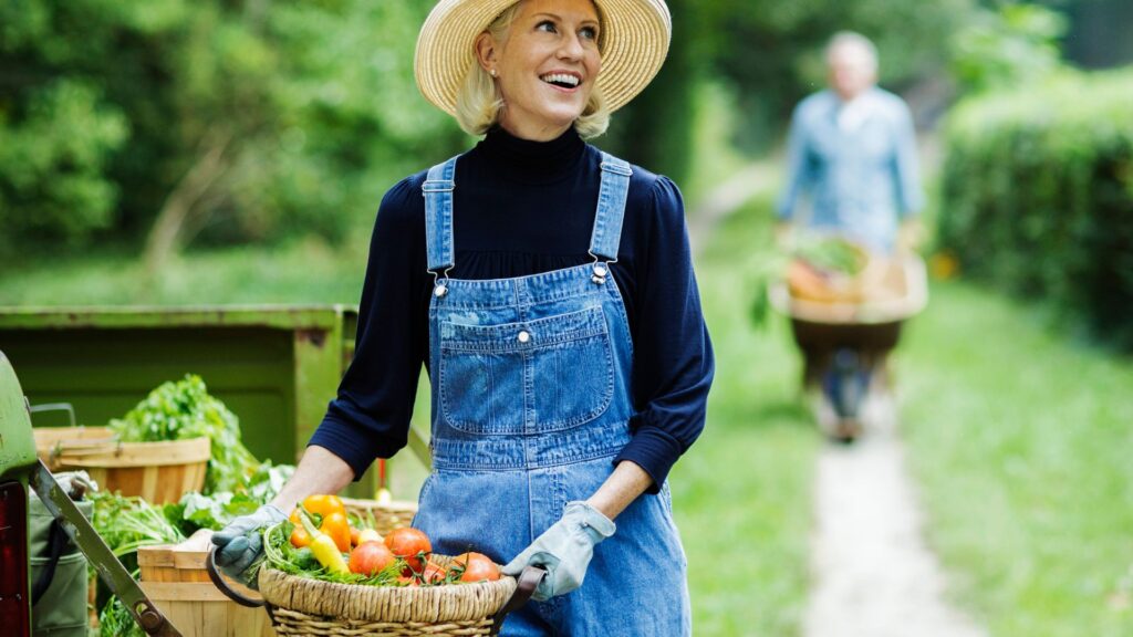 Mulher sorridente com chapéu de palha e jardineira jeans segurando uma cesta cheia de legumes frescos colhidos da horta.