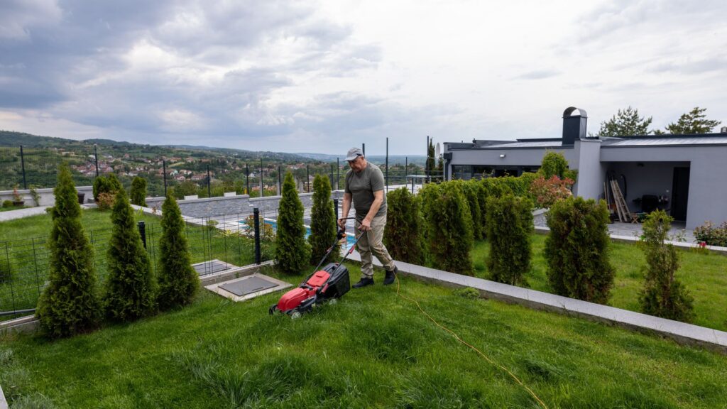 Homem utilizando um cortador de grama elétrico em um terreno residencial com inclinação e grama alta, ilustrando a escolha do cortador de grama para terrenos irregulares conforme o tamanho e tipo de terreno.