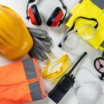 Equipamentos de proteção individual (EPI) sobre uma mesa branca: capacete amarelo, protetor auricular, luvas, óculos, respirador, colete refletivo e máscara, usados por operadores de roçadeira.