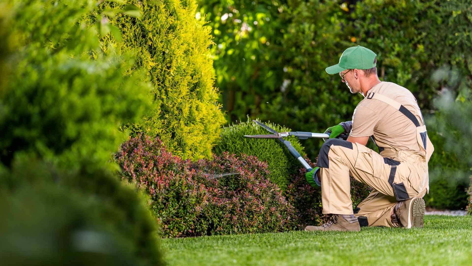Homem cuidando de jardim com tesoura de poda, usando equipamentos de proteção e ferramentas de jardinagem.