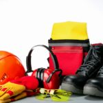 EPIs sobre superfície cinza: capacete laranja, luvas de segurança, protetor auricular, óculos de proteção, botas de segurança e colete refletivo — usados conforme as NRs 6, 12 e 31 para operadores de roçadeira.