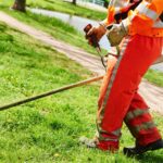 trabalhador usando roçadeira a gasolina para cortar grama alta em área verde próxima a um lago