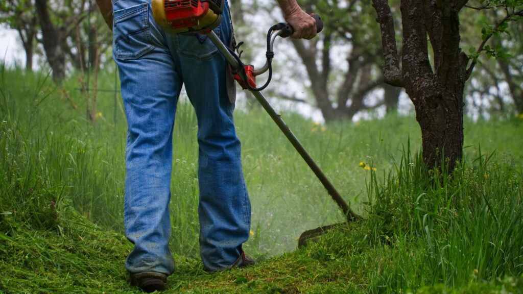 Homem utilizando roçadeira a gasolina para cortar grama alta em terreno com árvores