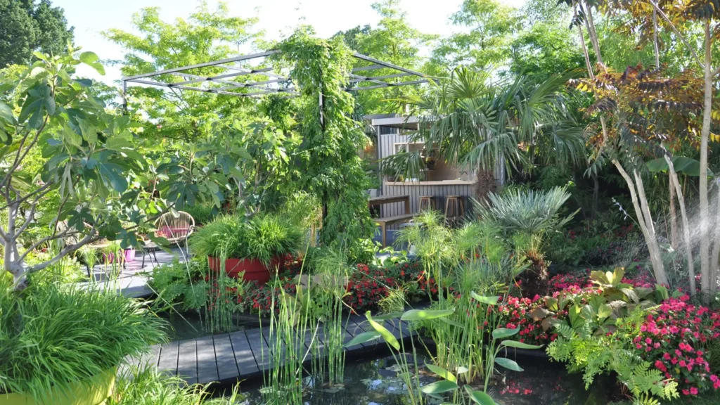 Jardim tropical exuberante com diversas plantas verdes, flores coloridas e um deck de madeira ao lado de um espelho d’água.