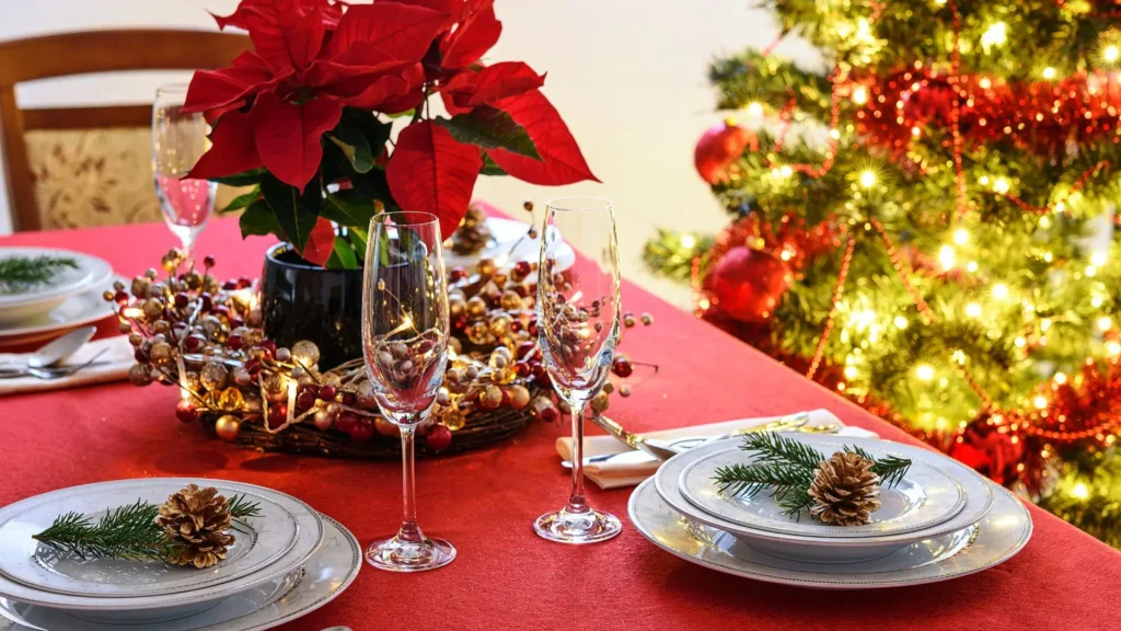 Flor de Natal vermelha decorando uma mesa de jantar posta para o Natal com louças, taças e árvore iluminada ao fundo.