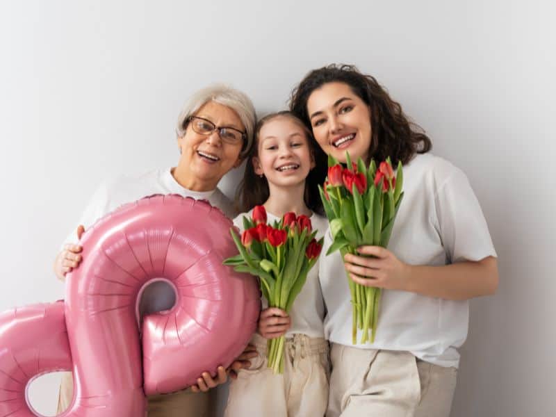 Mulheres de três gerações segurando flores, representando lembrancinhas do Dia da Mulher em contexto familiar