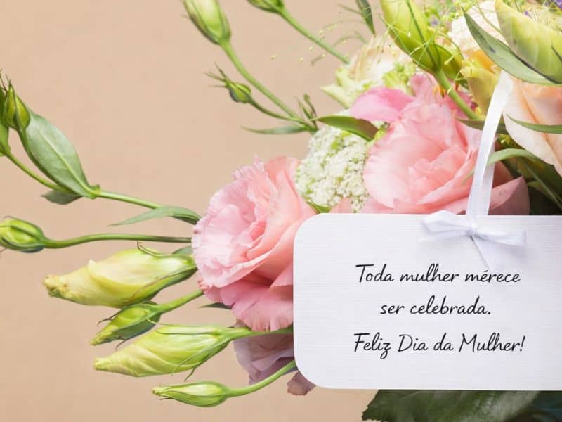 cartão com mensagem curta feliz dia da mulher pendurado em arranjo de flores rosadas representando mensagens dia da mulher curtas que funcionam em qualquer contexto