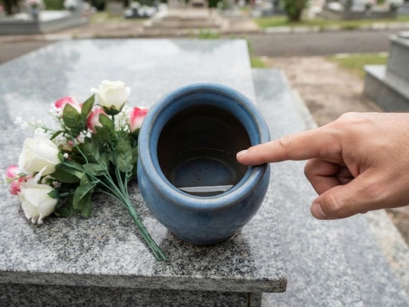 Mão apontando água acumulada dentro de um recipiente azul ao lado de flores artificiais no cemitério.