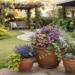 Trio de vasos com flores ornamentais em jardim residencial bem cuidado, com vaso maior em destaque e pergolado ao fundo.