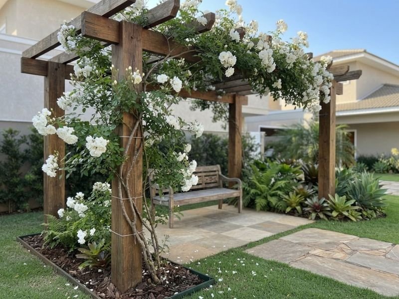 Roseira de flores brancas cobrindo parcialmente um pergolado de madeira com banco ao fundo.