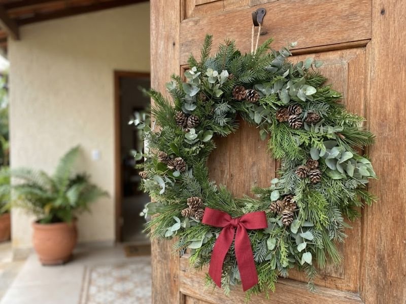 Guirlanda de folhagens verdes, eucalipto e pinhas com laço vermelho pendurada em uma porta de madeira.